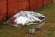A silver emergency blanket covers a person-shaped form on grass beside a red wooden wall, with crutches nearby, creating a somber scene.