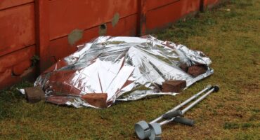 A silver emergency blanket covers a person-shaped form on grass beside a red wooden wall, with crutches nearby, creating a somber scene.