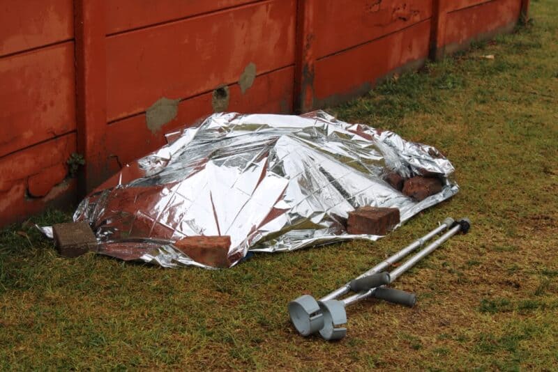 A silver emergency blanket covers a person-shaped form on grass beside a red wooden wall, with crutches nearby, creating a somber scene.