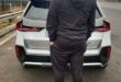 A man in a dark cap and tracksuit stands behind a white SUV on an overcast roadside, creating a quiet, contemplative mood.