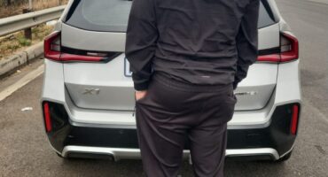 A man in a dark cap and tracksuit stands behind a white SUV on an overcast roadside, creating a quiet, contemplative mood.