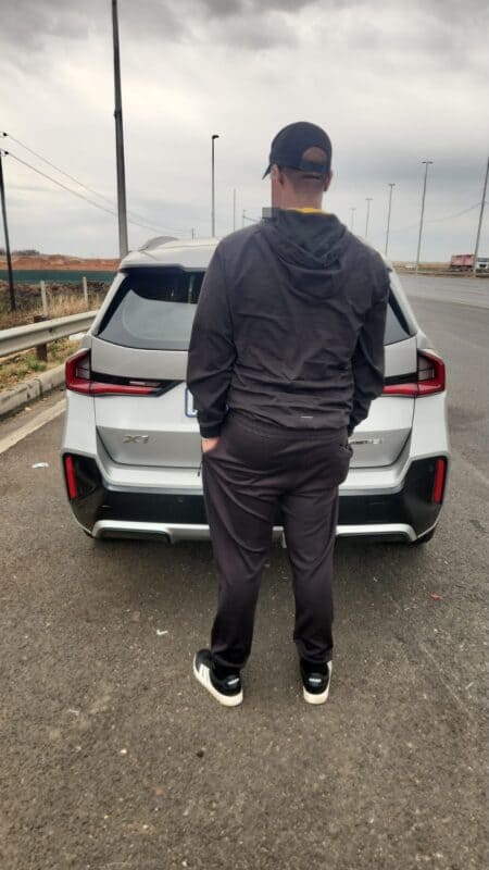 A man in a dark cap and tracksuit stands behind a white SUV on an overcast roadside, creating a quiet, contemplative mood.