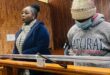 Solemn courtroom scene: a woman in a navy coat and scarf stands beside a masked person in a knit hat and gray hoodie behind the witness rail.