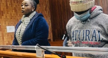 Solemn courtroom scene: a woman in a navy coat and scarf stands beside a masked person in a knit hat and gray hoodie behind the witness rail.