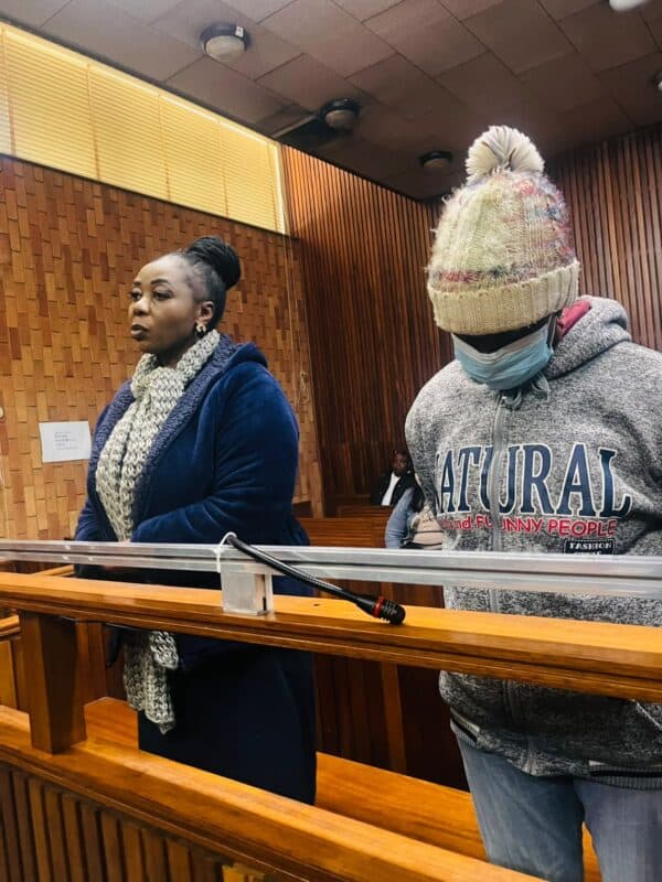 Solemn courtroom scene: a woman in a navy coat and scarf stands beside a masked person in a knit hat and gray hoodie behind the witness rail.