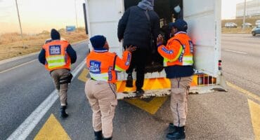 EMPD officers in orange safety vests help a bundled traveler into a white truck on a highway shoulder, creating a cooperative scene.