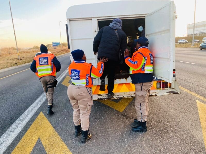 EMPD officers in orange safety vests help a bundled traveler into a white truck on a highway shoulder, creating a cooperative scene.