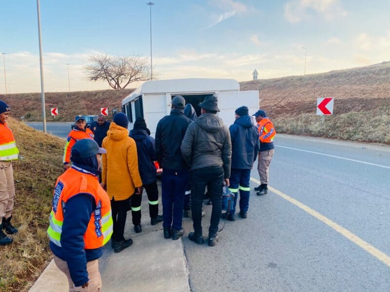 Roadside emergency crew in bright orange safety vests gathers around a white van on a curving winter road marked with red chevrons, focused.