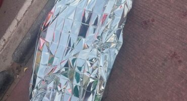 Person-shaped figure wrapped in a reflective silver emergency blanket lies beside a curb on a brick sidewalk, creating a somber, urgent scene.