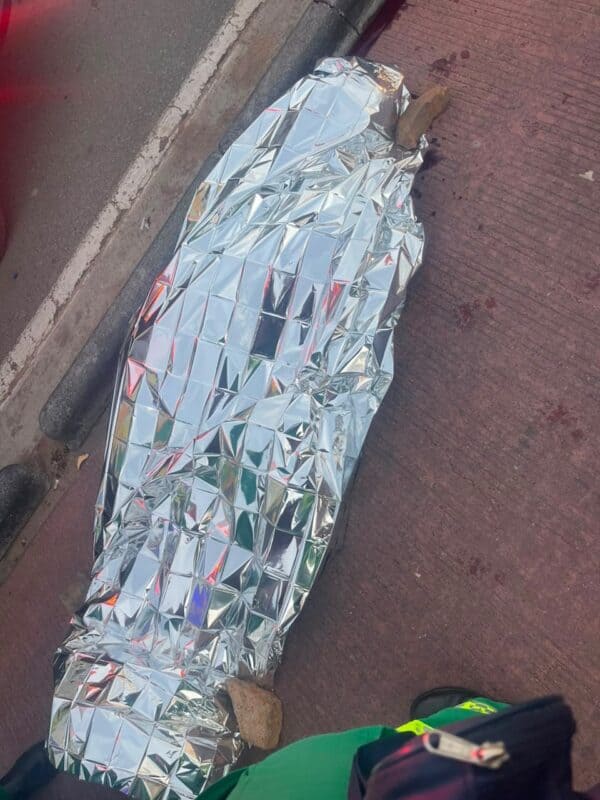Person-shaped figure wrapped in a reflective silver emergency blanket lies beside a curb on a brick sidewalk, creating a somber, urgent scene.