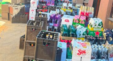 Colorful beverage crates and boxes of beer, soda, and bottled drinks crowd a small storefront, creating a busy, vibrant market scene.