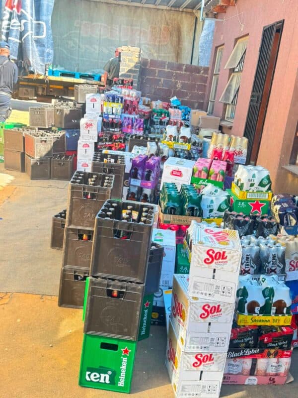 Colorful beverage crates and boxes of beer, soda, and bottled drinks crowd a small storefront, creating a busy, vibrant market scene.