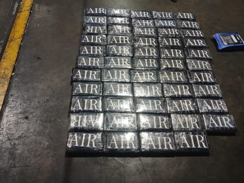 Rows of shiny rectangular blocks stamped “AIR” form a grid on a dark industrial floor beside a yellow safety line and blue device.