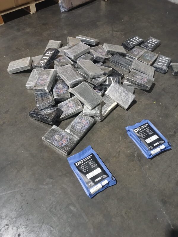 A pile of silver-wrapped rectangular packages lies on a concrete floor beside two blue DC-branded packages, creating an investigative tone.