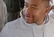 Black man in a gray hoodie holds a water bottle while seated among a crowd, looking to the side with a calm, thoughtful expression.