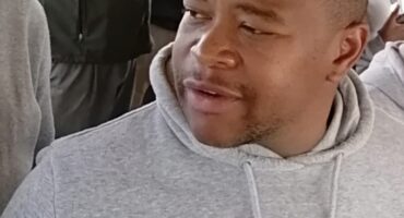 Black man in a gray hoodie holds a water bottle while seated among a crowd, looking to the side with a calm, thoughtful expression.