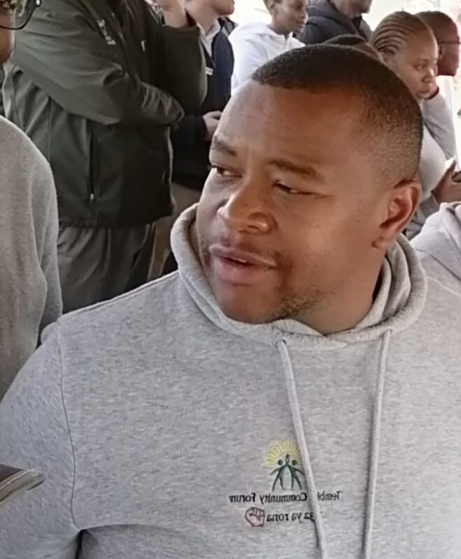 Black man in a gray hoodie holds a water bottle while seated among a crowd, looking to the side with a calm, thoughtful expression.