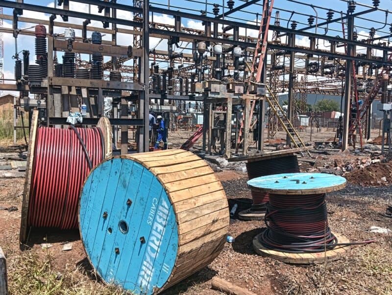 Large wooden cable reels, including blue and red spools, sit in a sunny electrical substation filled with transformers and power lines.