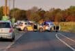 Police checkpoint blocks a rural road, with officers beside flashing patrol cars and stopped vehicles under warm evening light.