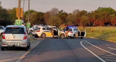 Police checkpoint blocks a rural road, with officers beside flashing patrol cars and stopped vehicles under warm evening light.