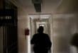 A silhouetted person walks down a dim institutional hallway past a barred office door and fire extinguisher, creating a tense, solitary mood.