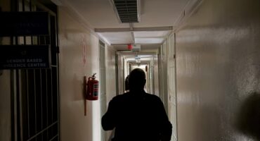 A silhouetted person walks down a dim institutional hallway past a barred office door and fire extinguisher, creating a tense, solitary mood.