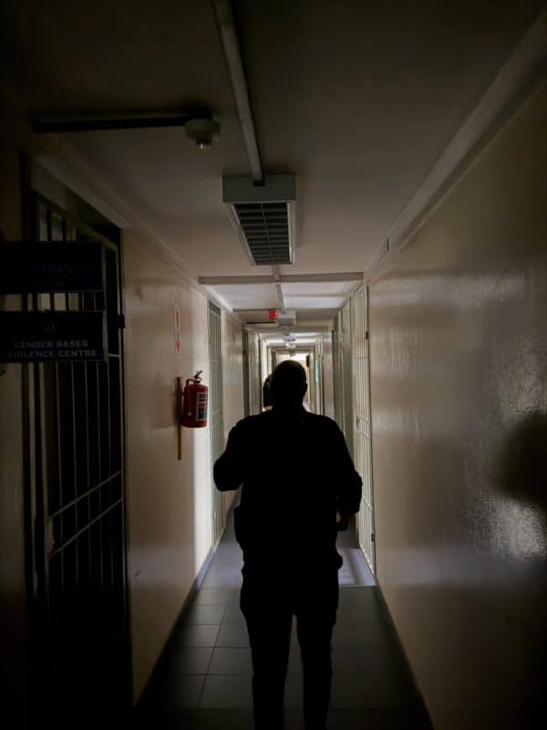 A silhouetted person walks down a dim institutional hallway past a barred office door and fire extinguisher, creating a tense, solitary mood.