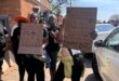 Two Black women hold handwritten anti-violence protest signs outside a brick building, surrounded by demonstrators and cars on a sunny day.