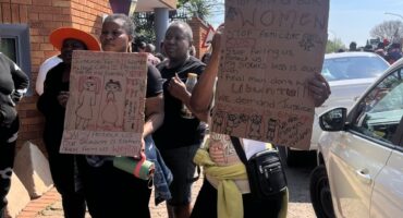 Two Black women hold handwritten anti-violence protest signs outside a brick building, surrounded by demonstrators and cars on a sunny day.
