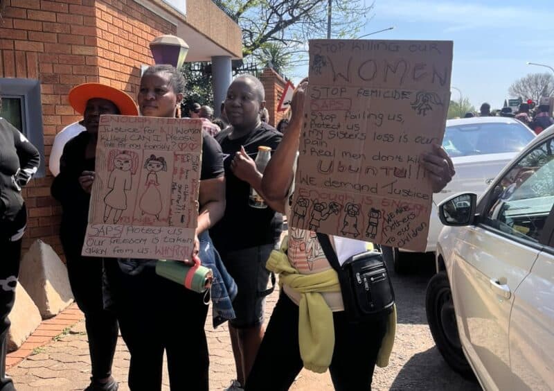 Two Black women hold handwritten anti-violence protest signs outside a brick building, surrounded by demonstrators and cars on a sunny day.