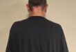Rear view of a balding man in an oversized black T-shirt and dark shorts, standing against a plain beige wall in a neutral, subdued setting.