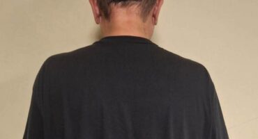 Rear view of a balding man in an oversized black T-shirt and dark shorts, standing against a plain beige wall in a neutral, subdued setting.