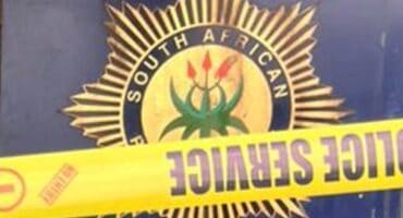 South African Police badge with a gold starburst emblem and national symbol, partially blocked by yellow “POLICE SEAL” crime scene tape.