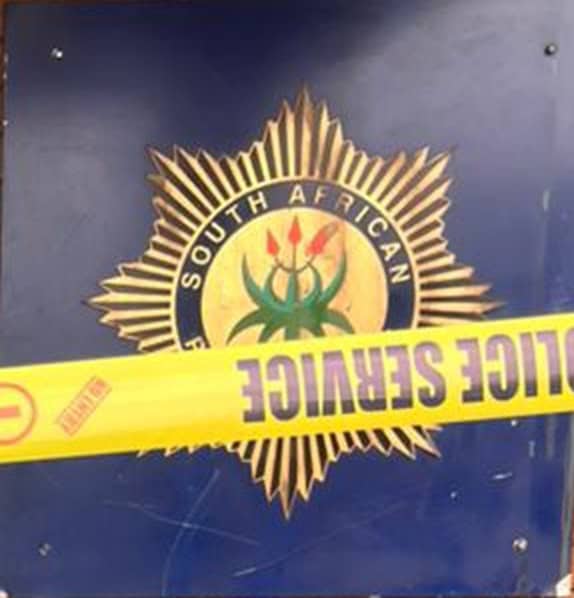 South African Police badge with a gold starburst emblem and national symbol, partially blocked by yellow “POLICE SEAL” crime scene tape.
