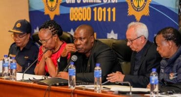 South African police officials sit at a press conference table beneath a SAPS Crime Stop banner, appearing serious and focused.