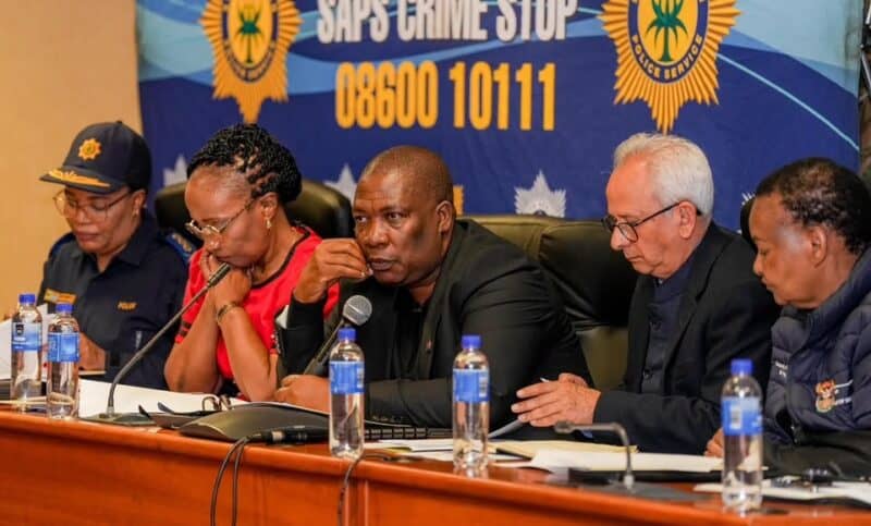 South African police officials sit at a press conference table beneath a SAPS Crime Stop banner, appearing serious and focused.