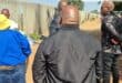 Men gather beside a tall concrete fence on a sunlit dirt path, listening to one another; their serious posture gives the outdoor meeting a tense tone.