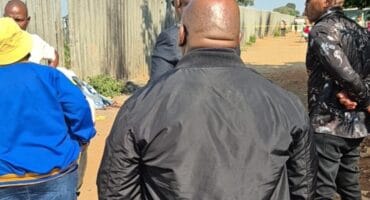 Men gather beside a tall concrete fence on a sunlit dirt path, listening to one another; their serious posture gives the outdoor meeting a tense tone.