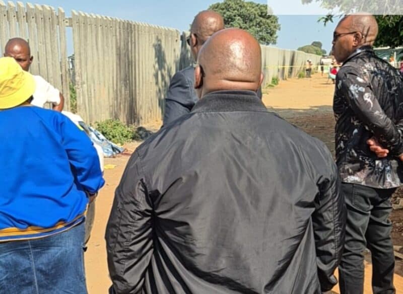 Men gather beside a tall concrete fence on a sunlit dirt path, listening to one another; their serious posture gives the outdoor meeting a tense tone.