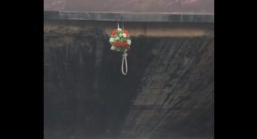 A small red-and-white flower arrangement with green foliage hangs by a loop beneath a dark, weathered concrete bridge, creating a quiet mood.