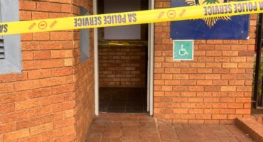 Brick police station entrance cordoned off with yellow SA Police Service tape; emblem, open doorway, and wheelchair sign create a tense scene.