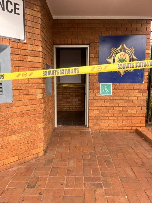 Brick police station entrance cordoned off with yellow SA Police Service tape; emblem, open doorway, and wheelchair sign create a tense scene.