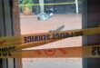 View through a doorway of a brick building entrance cordoned off with yellow police tape, scattered debris, potted plant, and damaged pavement.