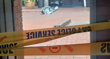 View through a doorway of a brick building entrance cordoned off with yellow police tape, scattered debris, potted plant, and damaged pavement.