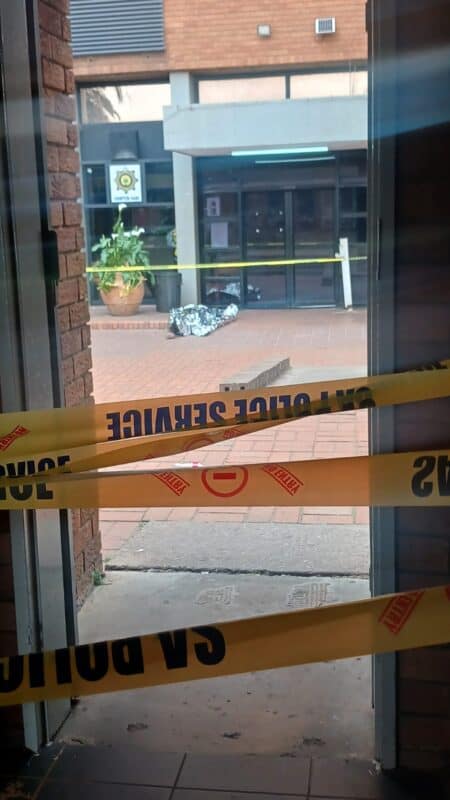 View through a doorway of a brick building entrance cordoned off with yellow police tape, scattered debris, potted plant, and damaged pavement.