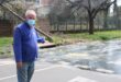 Masked man in a blue jacket gestures toward sewerage water rushing across a city street from an overflowing stream, conveying concern.