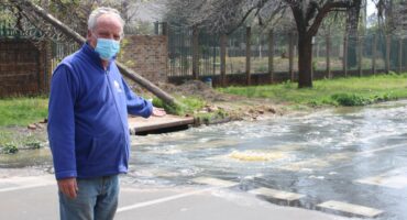 Masked man in a blue jacket gestures toward sewerage water rushing across a city street from an overflowing stream, conveying concern.