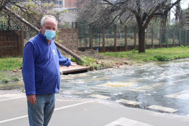 Masked man in a blue jacket gestures toward sewerage water rushing across a city street from an overflowing stream, conveying concern.