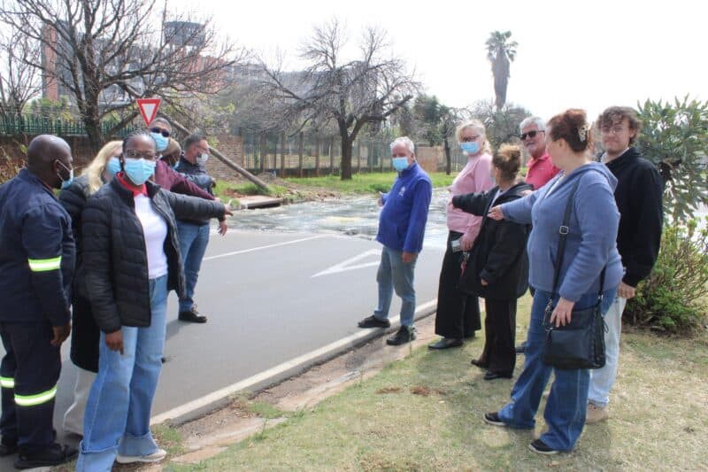 Masked community members gather beside a road with a safety worker, listening during an outdoor discussion on a cool, overcast day.