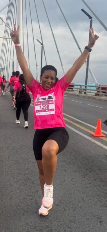 Smiling runner in a bright pink shirt celebrates mid-stride with arms raised in victory on a bridge, surrounded by runners and traffic cones.